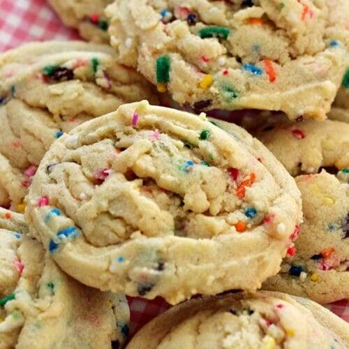 A dish lined with pink gingham parchment paper is topped with a pile of baked sprinkle cookies.