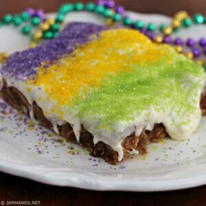 White plate with a serving of King Cake Bread Pudding with Bourbon Cream Cheese Frosting. It is topped with purple, yellow and green sprinkles.