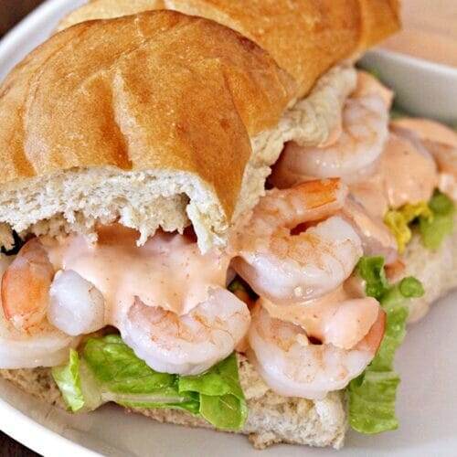 White plate with a French bread sandwich; filled with shrimp, zesty sauce, and lettuce.