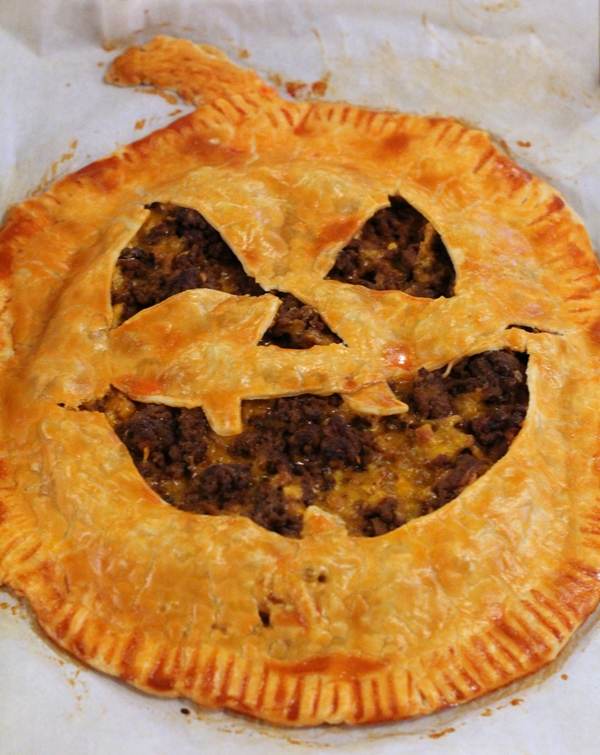 Close up of a pie crust brushed with an orange egg wash; shapes cut out of it to resemble a jack o' lantern for Halloween.