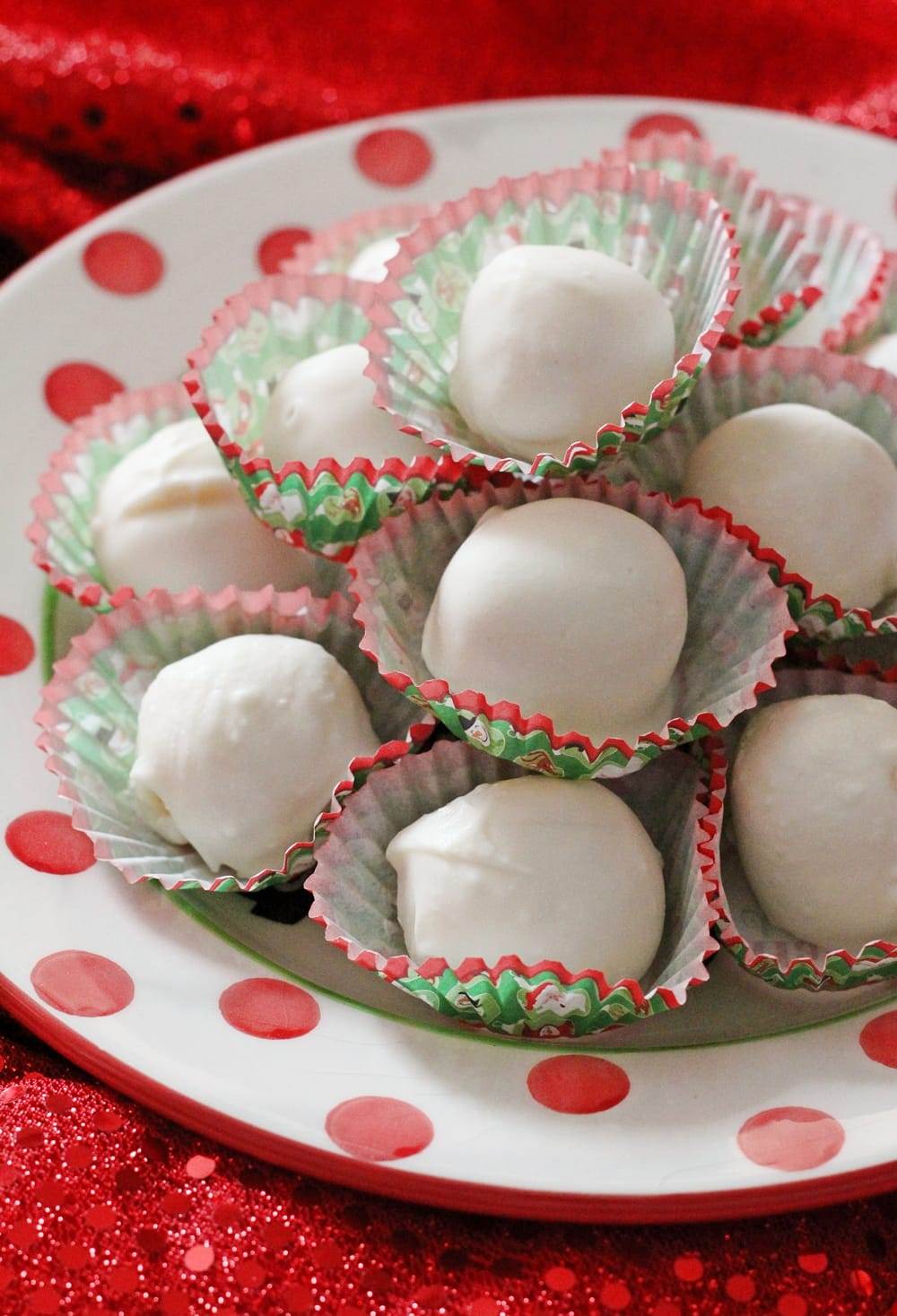 A red and white polka dot dish piled high with white eggnog truffles, each truffle in a red and green holiday candy liner.